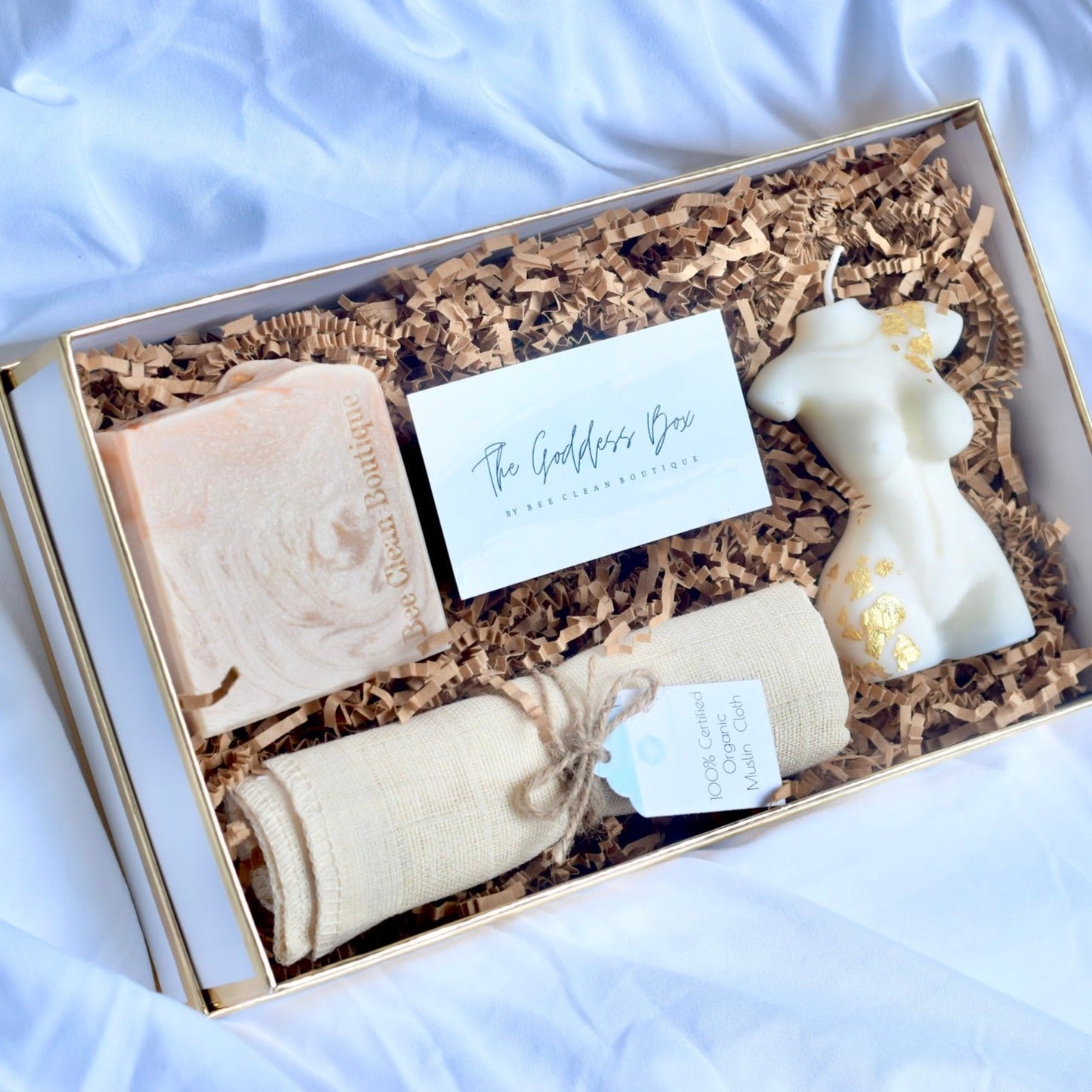 An open gift box showing a bar of artisanal soap, organic muslin cloth and a white body-shaped candle