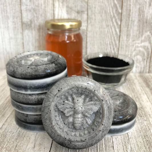 Five bars of charcoal facial soap with the bee icon on display in front of a jar of honey